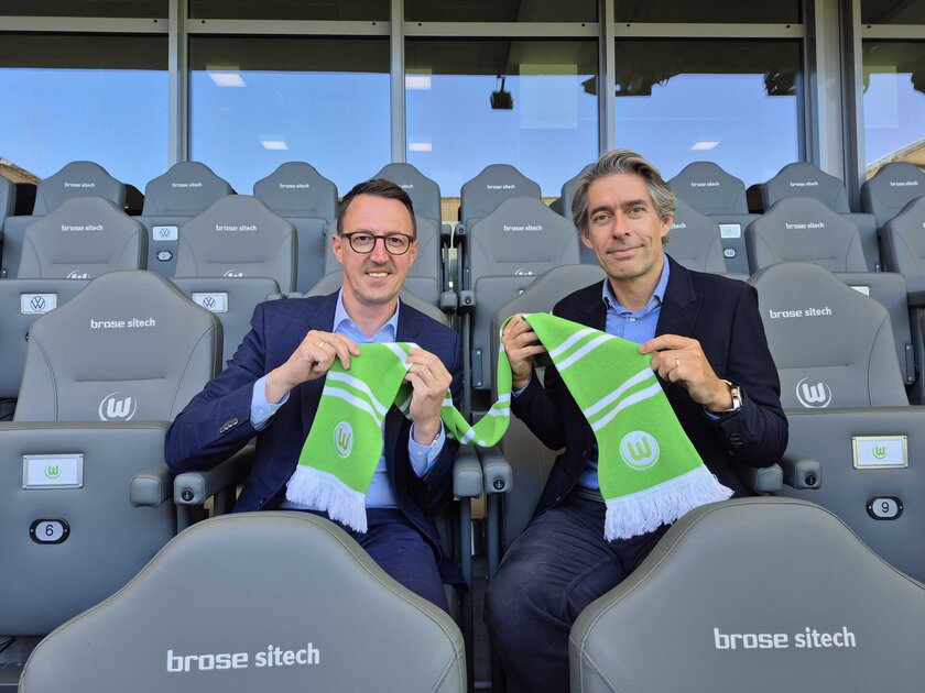 Michael Meeske, Geschäftsführung VfL Wolfsburg und Dalibor Dreznjak, Geschäftsführer der WOBCOM sitzen im Stadion und halten einen VfL-Wolfsburg Schal in die Kamera.